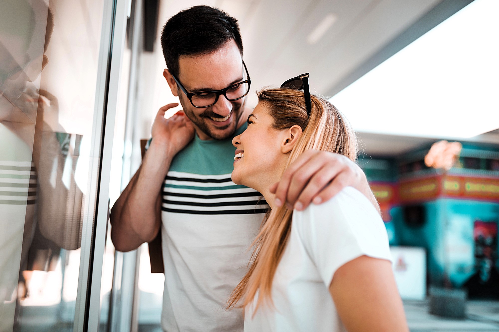 A man and woman are standing together outdoors, smiling and laughing. The man wears glasses and a striped shirt, gently wrapping his arm around the woman's shoulder. The woman, in a white shirt, looks up at him fondly. They are near a glass wall.