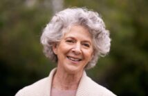 A smiling elderly woman with curly gray hair is wearing a beige sweater. She's outdoors, and a green, blurry background suggests trees or plants.