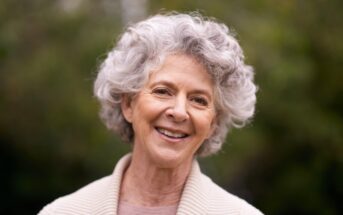 A smiling elderly woman with curly gray hair is wearing a beige sweater. She's outdoors, and a green, blurry background suggests trees or plants.