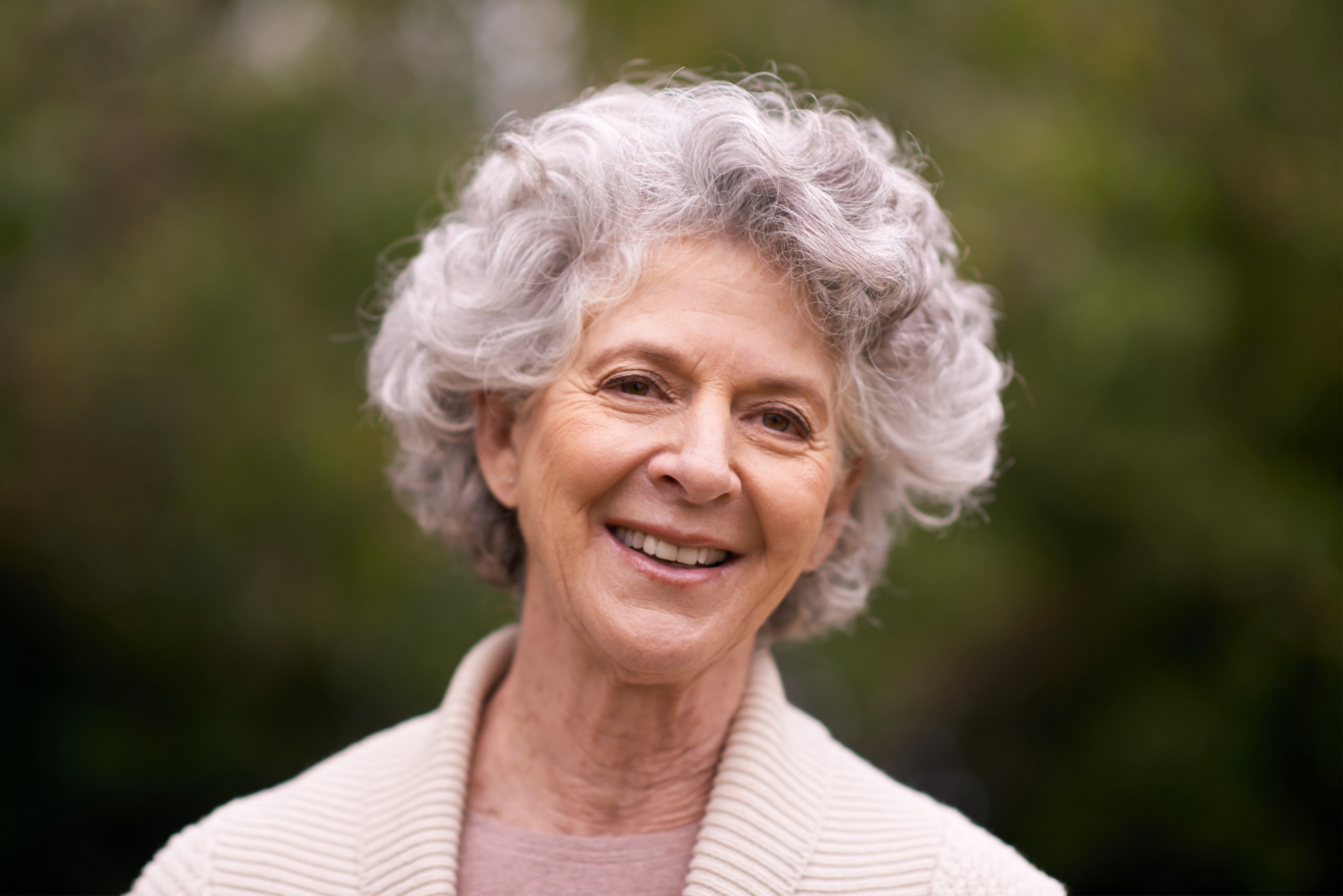 A smiling elderly woman with curly gray hair is wearing a beige sweater. She's outdoors, and a green, blurry background suggests trees or plants.