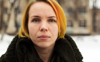 A person with short, yellow and orange hair stands outdoors in a snowy setting. They are wearing a dark scarf and have a neutral expression. The background is blurred with trees and buildings.