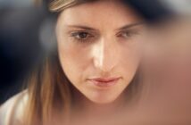 A close-up of a woman with long brown hair looking downward in a contemplative manner. Her expression appears thoughtful, with soft lighting highlighting her face. The background is blurred, focusing attention on her facial features.