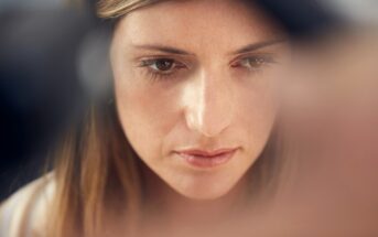 A close-up of a woman with long brown hair looking downward in a contemplative manner. Her expression appears thoughtful, with soft lighting highlighting her face. The background is blurred, focusing attention on her facial features.