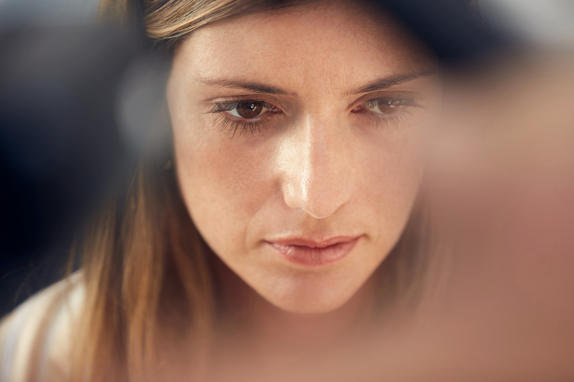 A close-up of a woman with long brown hair looking downward in a contemplative manner. Her expression appears thoughtful, with soft lighting highlighting her face. The background is blurred, focusing attention on her facial features.