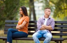 A woman and a man sit on a park bench with back-to-back posture, arms crossed, looking away from each other, suggesting disagreement or tension. The background shows blurred trees, indicating an outdoor setting.