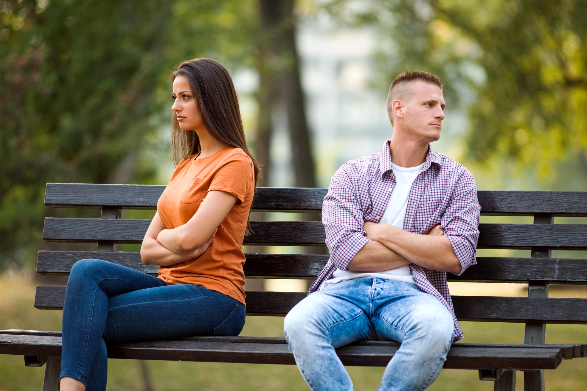 A woman and a man sit on a park bench with back-to-back posture, arms crossed, looking away from each other, suggesting disagreement or tension. The background shows blurred trees, indicating an outdoor setting.