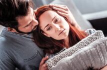 A man gently comforts a sad woman by resting his head on hers, while she leans against a grey knitted pillow. They are sitting close together on a couch, with a soft-focus background.