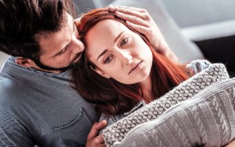 A man gently comforts a sad woman by resting his head on hers, while she leans against a grey knitted pillow. They are sitting close together on a couch, with a soft-focus background.