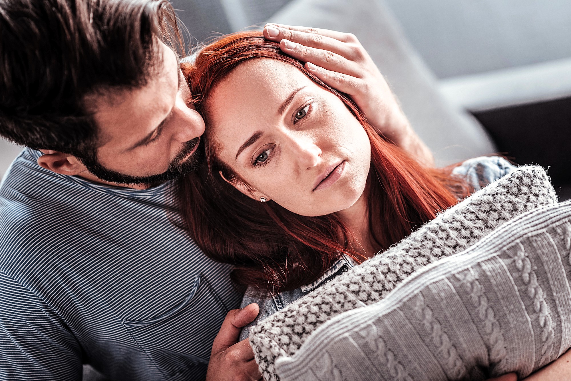 A man gently comforts a sad woman by resting his head on hers, while she leans against a grey knitted pillow. They are sitting close together on a couch, with a soft-focus background.