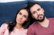 A woman and a man are sitting close together on a blue couch. Both have dark hair and are wearing casual tops, the woman in pink and the man in maroon. They are looking directly at the camera with neutral expressions.