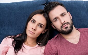 A woman and a man are sitting close together on a blue couch. Both have dark hair and are wearing casual tops, the woman in pink and the man in maroon. They are looking directly at the camera with neutral expressions.