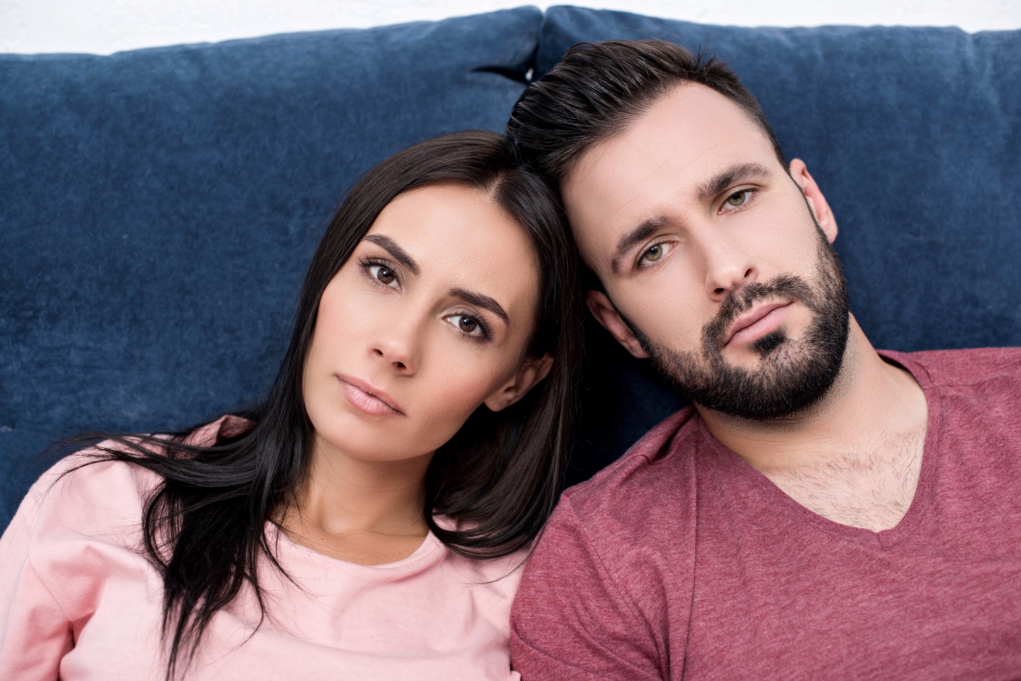 A woman and a man are sitting close together on a blue couch. Both have dark hair and are wearing casual tops, the woman in pink and the man in maroon. They are looking directly at the camera with neutral expressions.