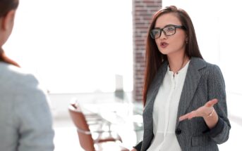 A woman with long brown hair and glasses is speaking passionately. She is wearing a gray blazer and white shirt, gesturing with one hand. Office chairs and a bright window are in the background, suggesting a professional setting.