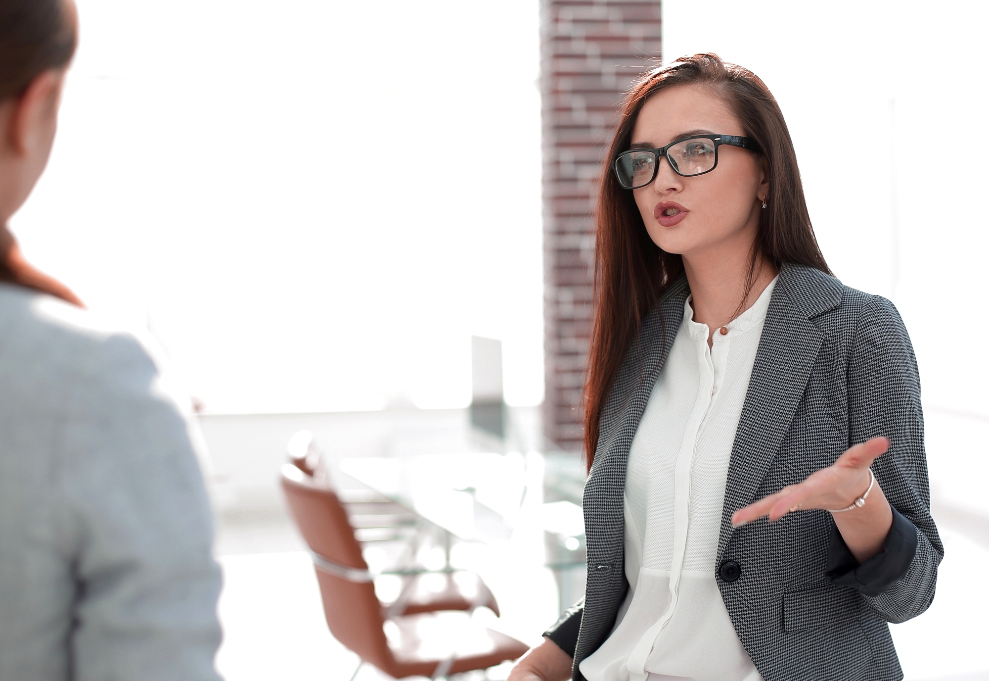 A woman with long brown hair and glasses is speaking passionately. She is wearing a gray blazer and white shirt, gesturing with one hand. Office chairs and a bright window are in the background, suggesting a professional setting.