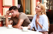 A woman and a man sit at an outdoor café table with cups of coffee. The woman appears to be talking and smiling, with her hand on her chest. The man looks pensive, resting his head on his hand. The setting is casual and sunny.