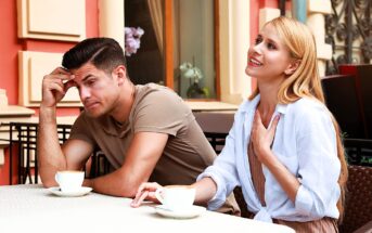 A woman and a man sit at an outdoor café table with cups of coffee. The woman appears to be talking and smiling, with her hand on her chest. The man looks pensive, resting his head on his hand. The setting is casual and sunny.