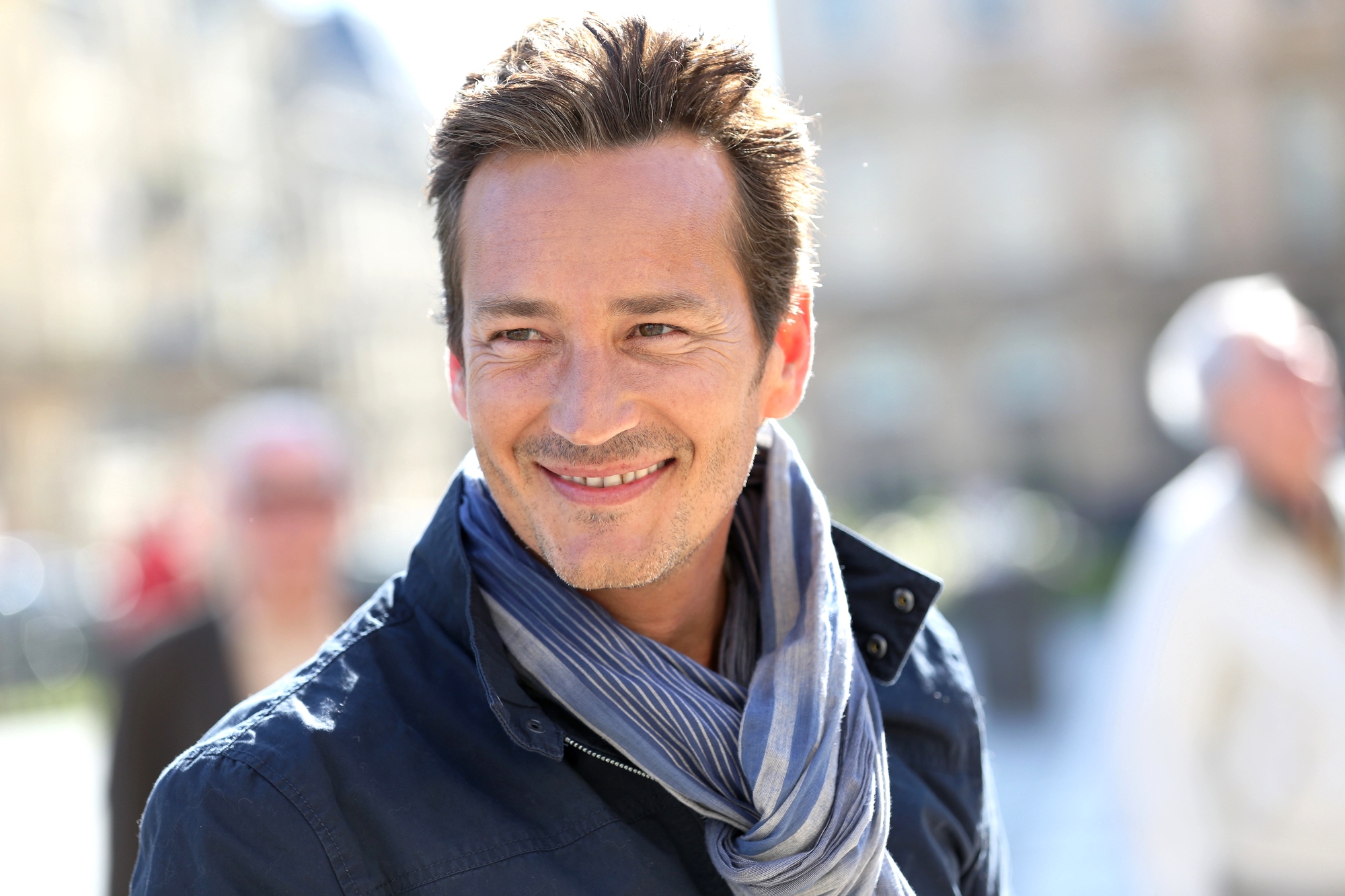 A man with short hair smiles outdoors. He is wearing a dark jacket and a light blue scarf. The background is blurred, showing people and architecture in soft focus on a sunny day.