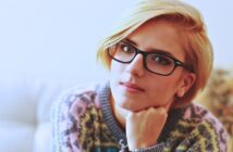 A person with short blond hair and black-rimmed glasses is gazing intently at the camera. They're wearing a colorful patterned sweater and resting their chin on their hand. The background is softly blurred.