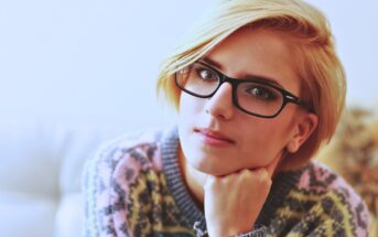 A person with short blond hair and black-rimmed glasses is gazing intently at the camera. They're wearing a colorful patterned sweater and resting their chin on their hand. The background is softly blurred.