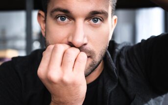 A close-up of a man with short hair and facial stubble, looking into the camera. He is wearing a dark jacket and has his hand partially covering his mouth, conveying a thoughtful or pensive expression.