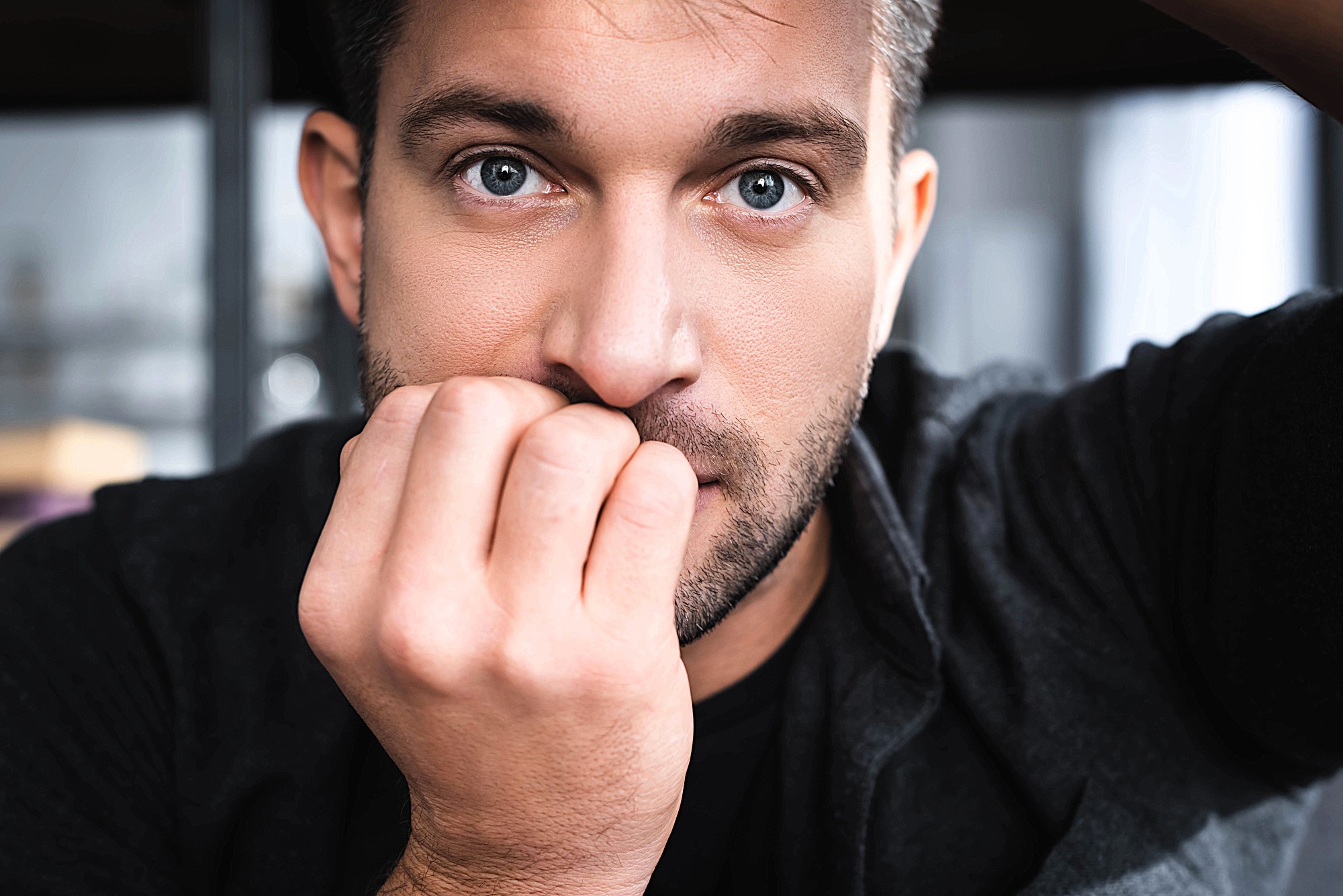 A close-up of a man with short hair and facial stubble, looking into the camera. He is wearing a dark jacket and has his hand partially covering his mouth, conveying a thoughtful or pensive expression.