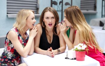 Three women sitting at a table outdoors. The woman in the center looks surprised, while the two on either side are whispering in her ears. There is a red flower pot on the table.