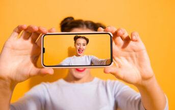 A person holds a smartphone in front of their face, capturing a selfie. The phone displays their excited expression with a wide smile. The background is a vibrant yellow, and the person wears a light shirt with their hair styled in two buns.