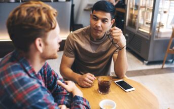 Two men sit at a wooden table in a café. One holds glasses, while the other listens attentively. A smartphone and a glass of iced coffee are on the table. Shelves with mugs are visible in the background.