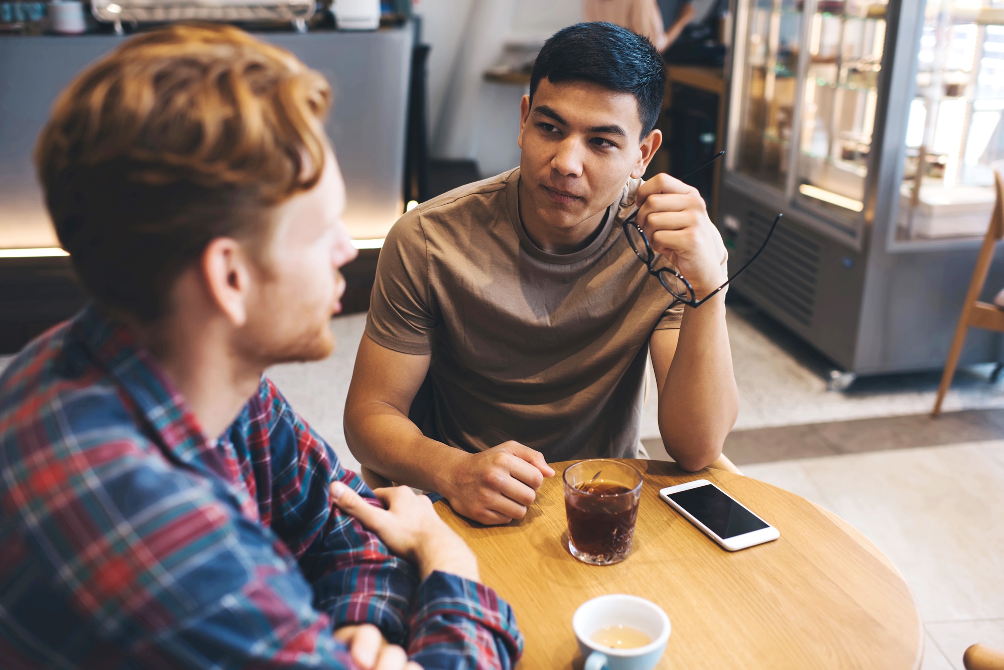 Two men sit at a wooden table in a café. One holds glasses, while the other listens attentively. A smartphone and a glass of iced coffee are on the table. Shelves with mugs are visible in the background.