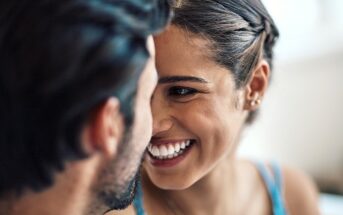 A close-up of a smiling couple facing each other. The woman, with braided hair and small earrings, beams while looking at the man, who has dark hair and a beard. The image captures a moment of affection and happiness.