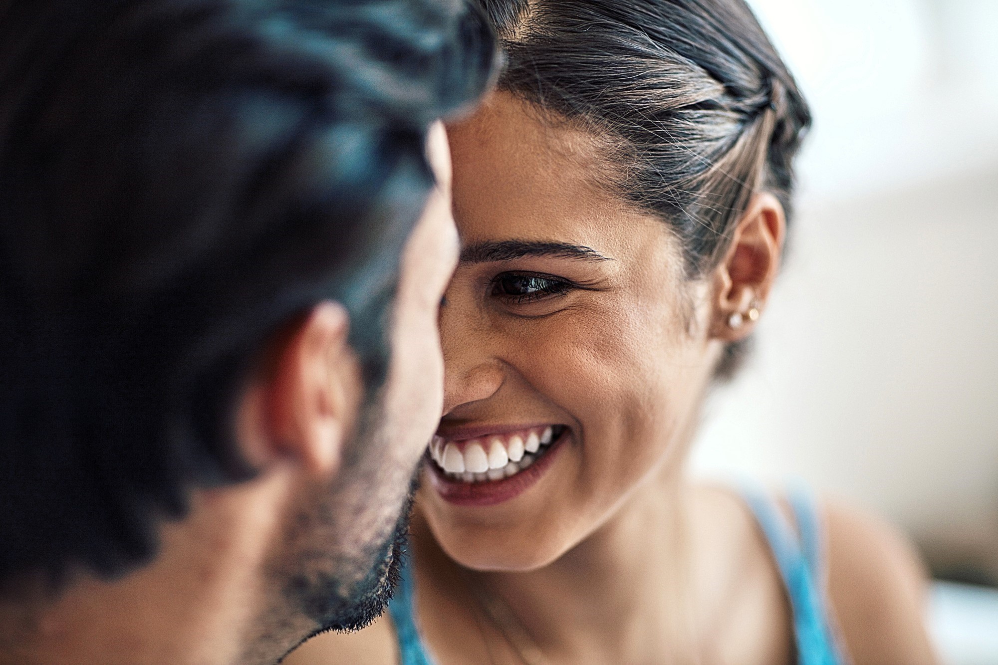 A close-up of a smiling couple facing each other. The woman, with braided hair and small earrings, beams while looking at the man, who has dark hair and a beard. The image captures a moment of affection and happiness.