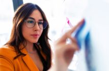 A woman with long brown hair and glasses is wearing a mustard yellow blazer. She is focused on writing or drawing on a clear board with colored markers in an office setting.