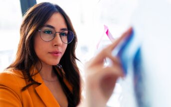 A woman with long brown hair and glasses is wearing a mustard yellow blazer. She is focused on writing or drawing on a clear board with colored markers in an office setting.