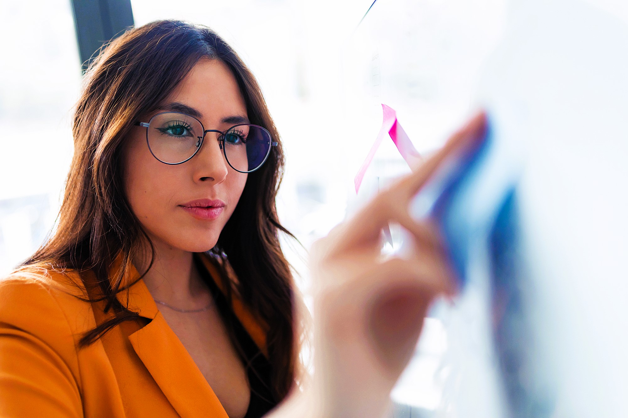 A woman with long brown hair and glasses is wearing a mustard yellow blazer. She is focused on writing or drawing on a clear board with colored markers in an office setting.