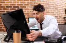 A man in an office setting leans forward in shock, staring at a computer monitor. He wears a white shirt and has a surprised expression. A coffee cup sits on the desk in front of him, and the wall behind is brick.