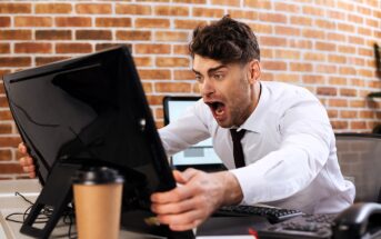 A man in an office setting leans forward in shock, staring at a computer monitor. He wears a white shirt and has a surprised expression. A coffee cup sits on the desk in front of him, and the wall behind is brick.