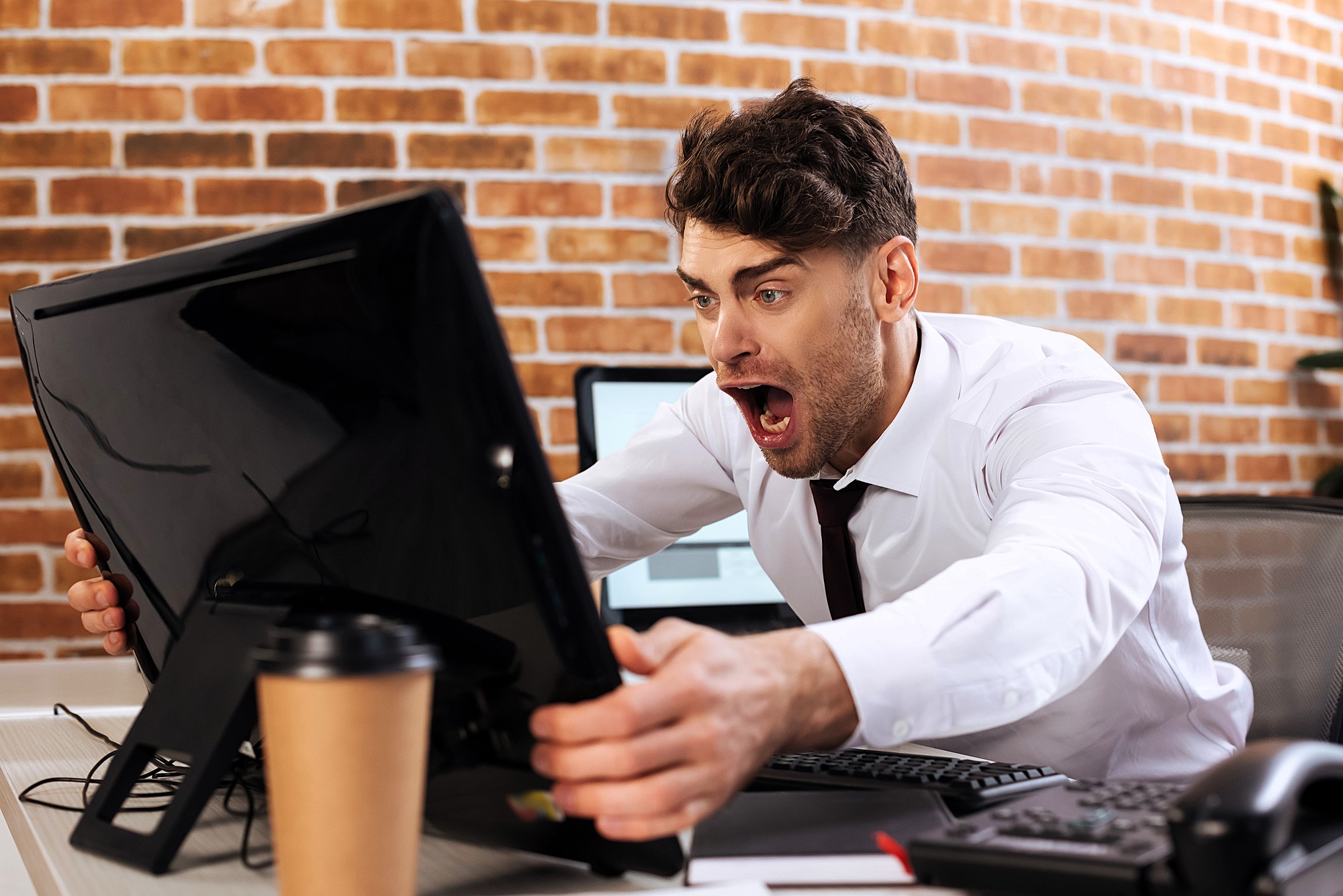 A man in an office setting leans forward in shock, staring at a computer monitor. He wears a white shirt and has a surprised expression. A coffee cup sits on the desk in front of him, and the wall behind is brick.