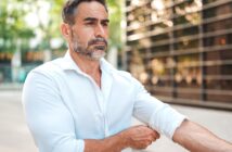 A man with a beard is adjusting the sleeve of his white shirt outdoors. He appears focused and is standing in front of a modern building with glass reflections. Trees are in the background, adding a touch of nature to the urban setting.