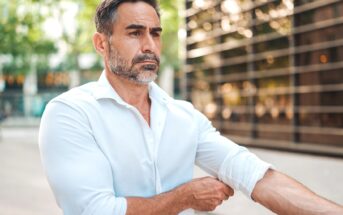 A man with a beard is adjusting the sleeve of his white shirt outdoors. He appears focused and is standing in front of a modern building with glass reflections. Trees are in the background, adding a touch of nature to the urban setting.