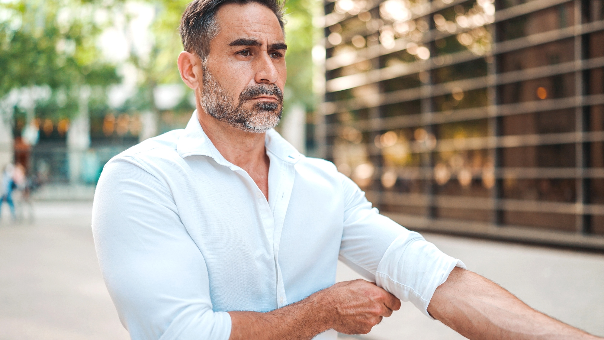 A man with a beard is adjusting the sleeve of his white shirt outdoors. He appears focused and is standing in front of a modern building with glass reflections. Trees are in the background, adding a touch of nature to the urban setting.