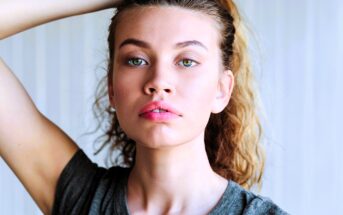 A person with wavy, light brown hair pulled back gazes directly at the camera. They are wearing a gray T-shirt and have an arm raised, touching their hair. The background is softly blurred.