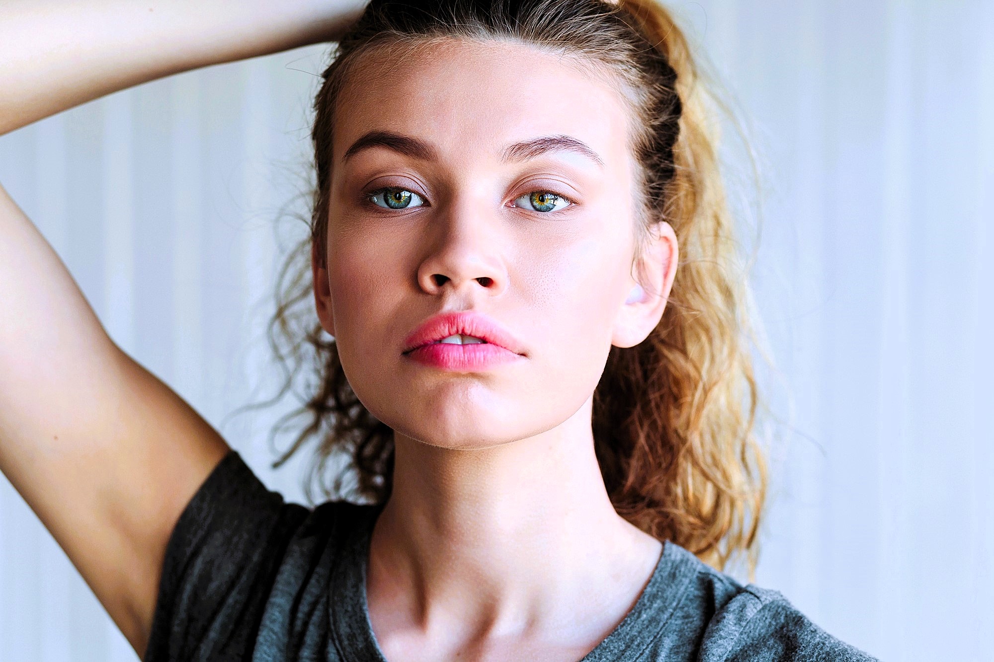 A person with wavy, light brown hair pulled back gazes directly at the camera. They are wearing a gray T-shirt and have an arm raised, touching their hair. The background is softly blurred.