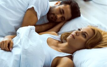 A man and woman are lying in bed under white sheets. The woman is awake, looking thoughtful, while the man is sleeping next to her. They are both wearing white tops, and the scene has a calm and serene atmosphere.