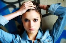 A woman with long hair, wearing a denim jacket, holds her hands on her head. She stands against a wall with artistic graffiti, looking directly at the camera with an intense expression.