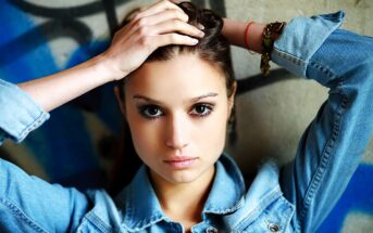 A woman with long hair, wearing a denim jacket, holds her hands on her head. She stands against a wall with artistic graffiti, looking directly at the camera with an intense expression.