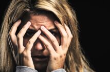 A person with blonde hair holds their hands over their face, peering through their fingers with a look of distress. The background is dark, emphasizing their expression.