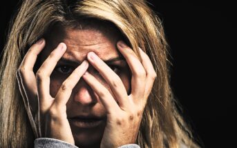 A person with blonde hair holds their hands over their face, peering through their fingers with a look of distress. The background is dark, emphasizing their expression.