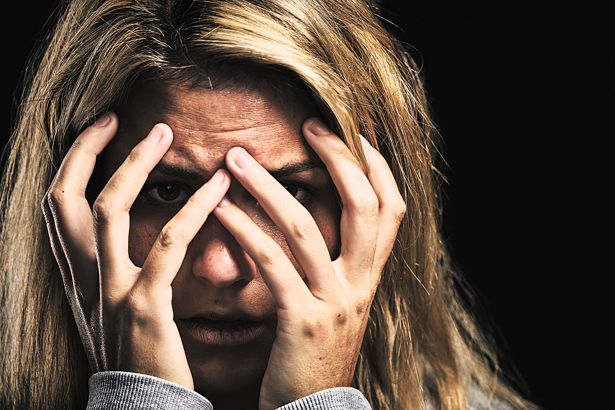 A person with blonde hair holds their hands over their face, peering through their fingers with a look of distress. The background is dark, emphasizing their expression.