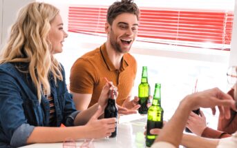 People are sitting around a table, smiling and talking while holding bottles of soda. Some wear casual outfits. The setting is bright, with red blinds in the background, suggesting a cheerful atmosphere.