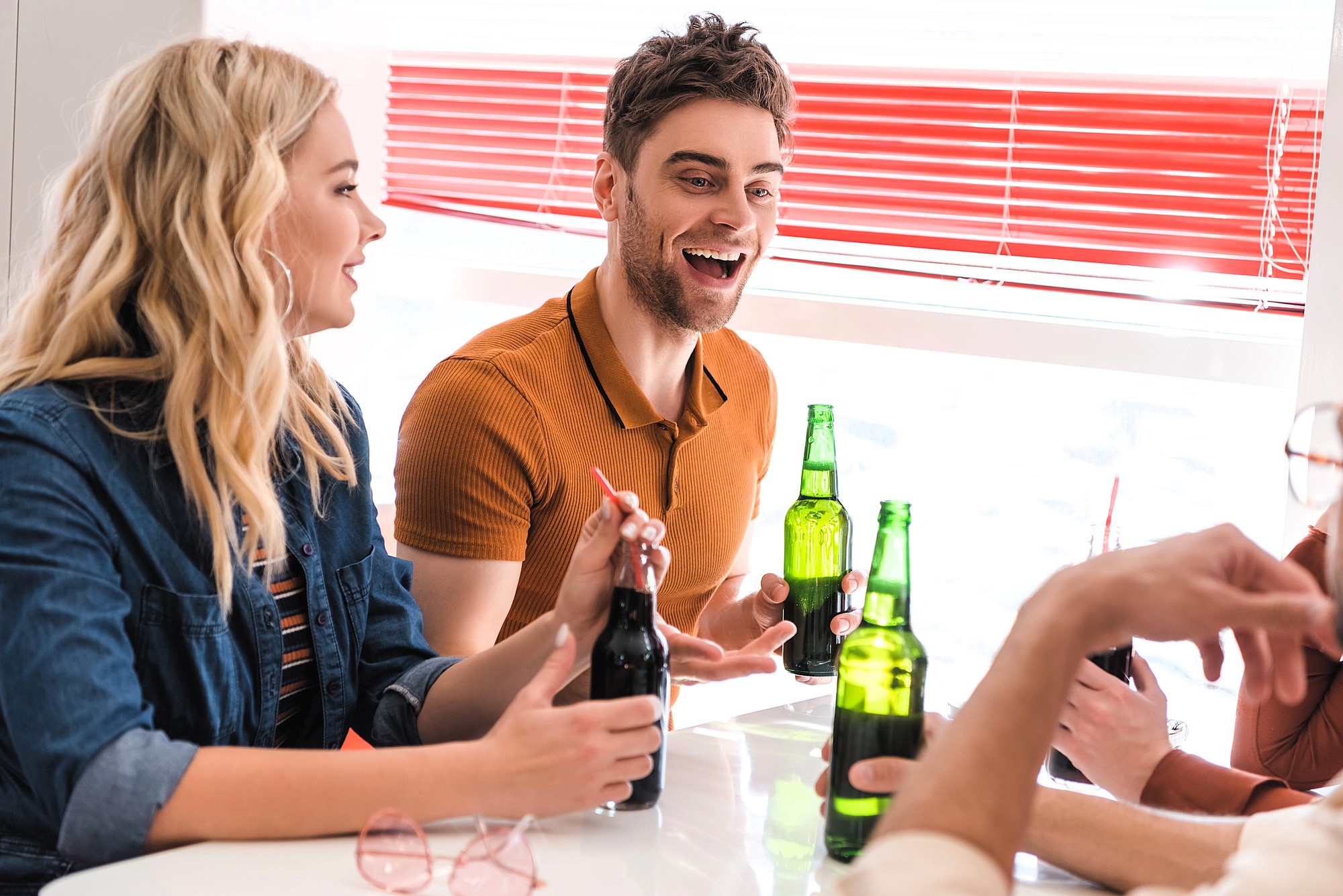 People are sitting around a table, smiling and talking while holding bottles of soda. Some wear casual outfits. The setting is bright, with red blinds in the background, suggesting a cheerful atmosphere.